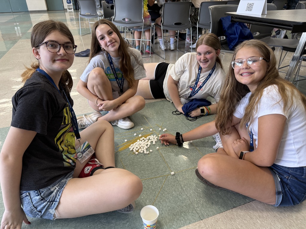 Four individuals are seated on the floor in a circle, participating in an activity where they build structures using uncooked spaghetti and marshmallows