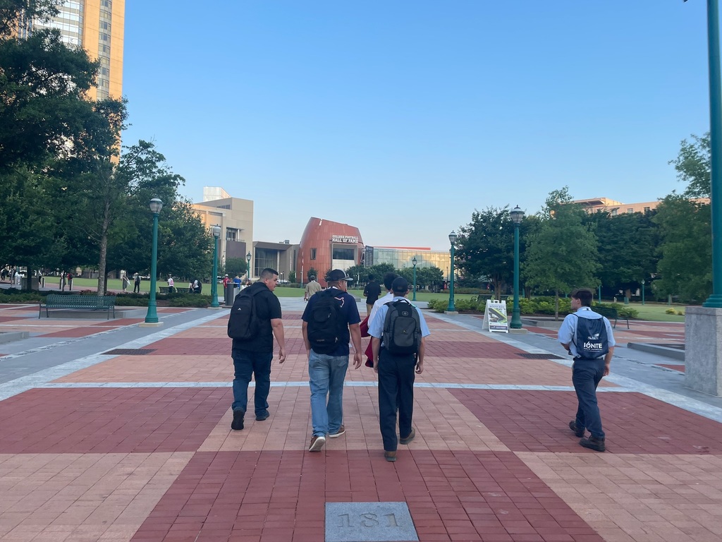 Five people walk on a paved pathway with red and beige tiles, carrying backpacks and heading toward a building in the distance. The area is surrounded by green trees, lamp posts, and benches.
