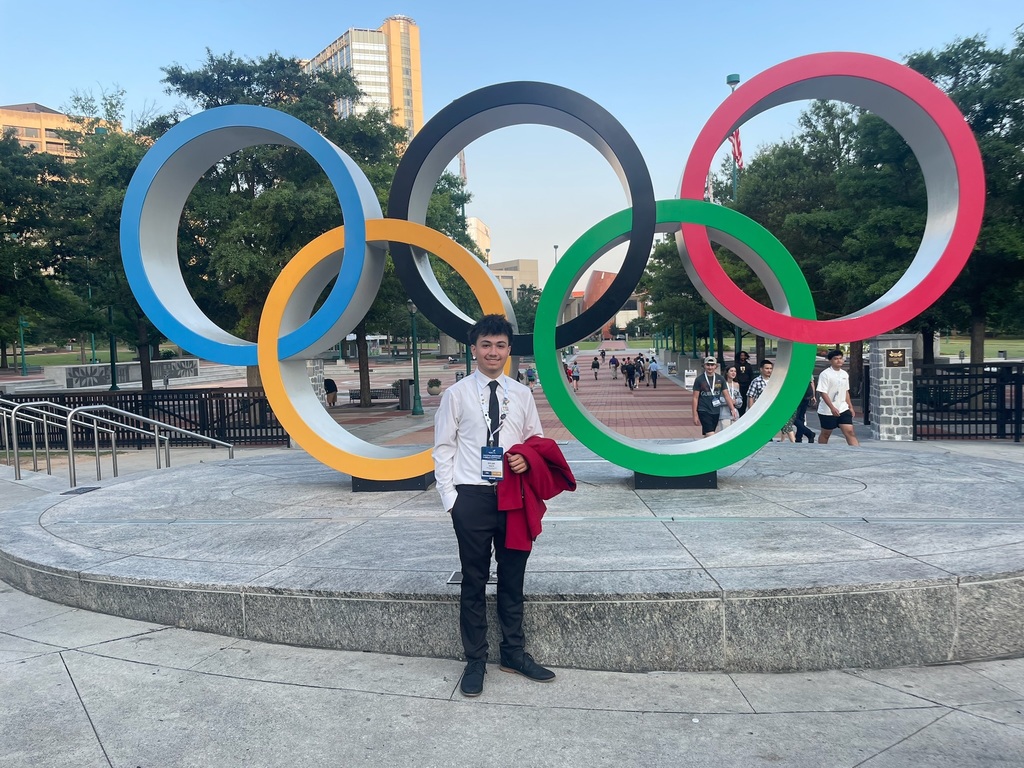 A person stands in front of a large sculpture of the Olympic rings, which are colored blue, yellow, black, green, and red. The person is dressed formally in a white shirt and black pants, holding a red jacket. Trees and buildings are visible in the background, with several other people walking around the area.