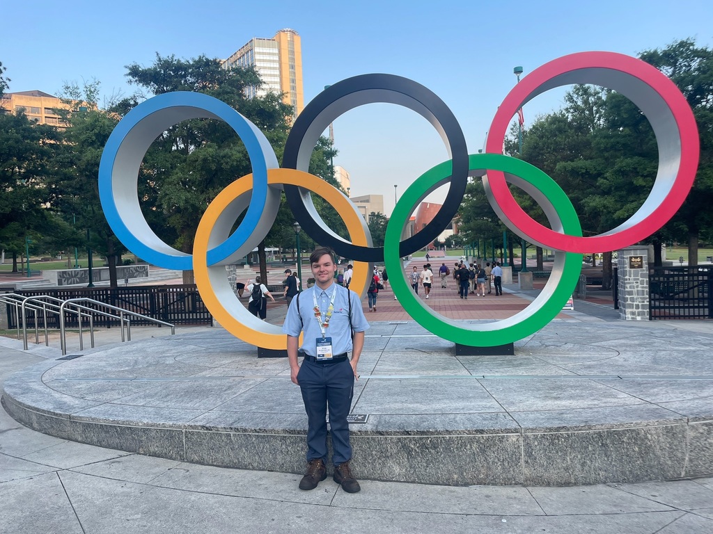 A person stands in front of a large sculpture of the Olympic rings, which are colored blue, yellow, black, green, and red. The sculpture is located in an outdoor plaza with trees and buildings visible in the background. Several people can be seen walking around the plaza. The person is wearing a light blue shirt and dark pants with a lanyard around their neck.