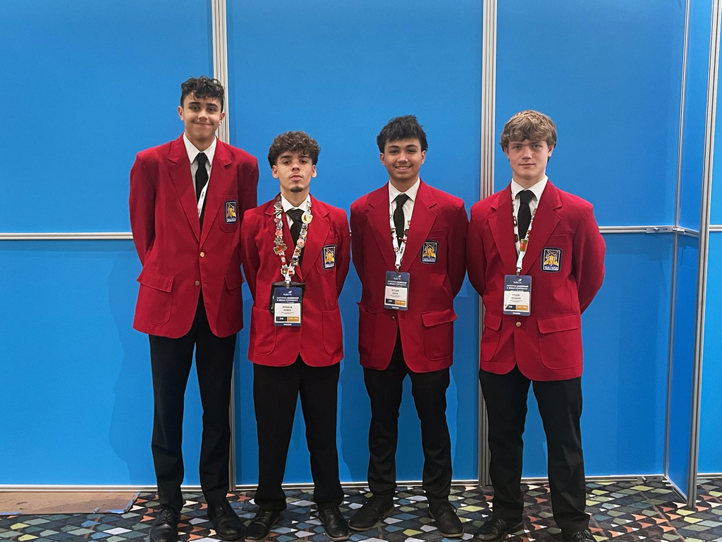 Four individuals stand side by side in front of a blue background. They are all wearing red blazers with a patch on the left chest, white shirts, black ties, and black pants. Each person has a name badge and lanyard around their neck. The floor is covered with a patterned carpet.