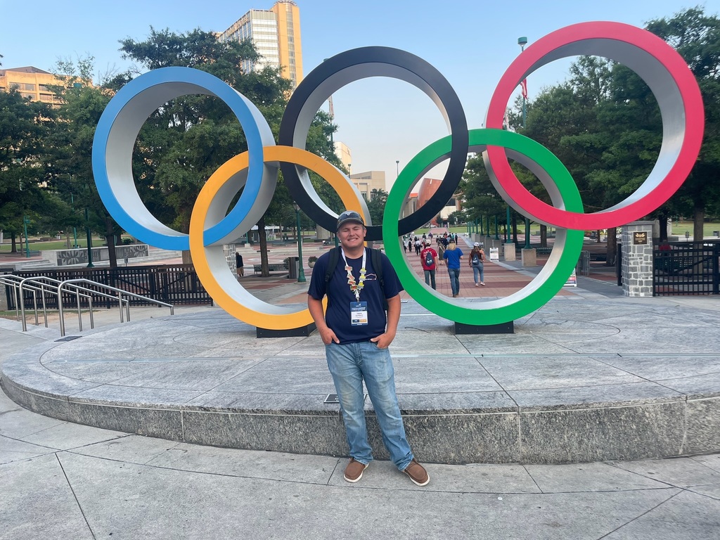 A person stands in front of a large sculpture of the Olympic rings, which are colored blue, yellow, black, green, and red. The background includes trees, buildings, and several other people walking in the area.