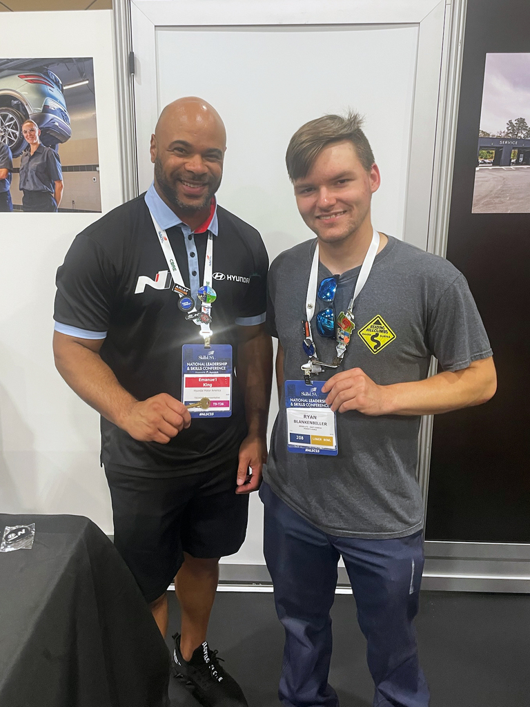 Two individuals stand side by side, both wearing lanyards with identification badges. The person on the left wears a black shirt with 'N' and 'Hyundai' logos, and the person on the right wears a gray shirt with a yellow logo.