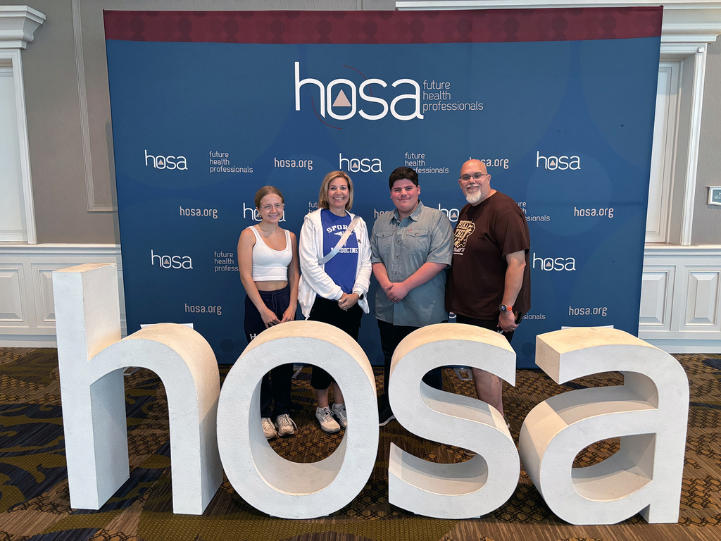 This image features four individuals standing in front of a backdrop that prominently displays the text "hosa future health professionals" and "hosa.org". In front of them are large 3D letters spelling out "hosa", indicating that the photo was taken at a HOSA event, which is associated with students interested in health professions.