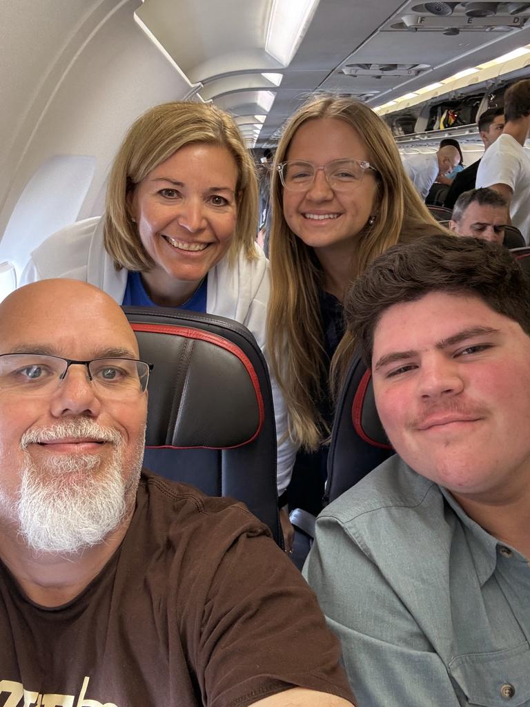 Four people inside an airplane. Two are seated in front, one wearing a brown shirt and the other a light blue shirt. Two others with long hair are standing behind them, leaning forward to join the photo. The airplane interior, including seats and overhead compartments, is visible.