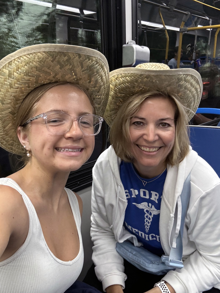 This image shows two individuals seated on a bus, both wearing straw hats. One is dressed in a white tank top and glasses, while the other is wearing a blue shirt that says "SPORT MEDICINE" with a medical symbol, and a white jacket. The setting appears casual, likely during a trip or outing, with the bus interior visible in the background.