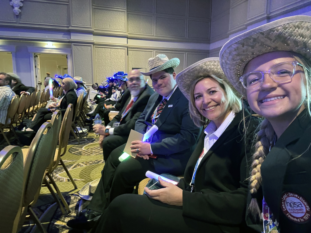 This image shows a group of people seated in rows, dressed in suits and wearing straw hats. Many are holding glowing sticks, and several have lanyards around their necks—suggesting they are attending a formal indoor event or conference, possibly with a celebratory or themed component.