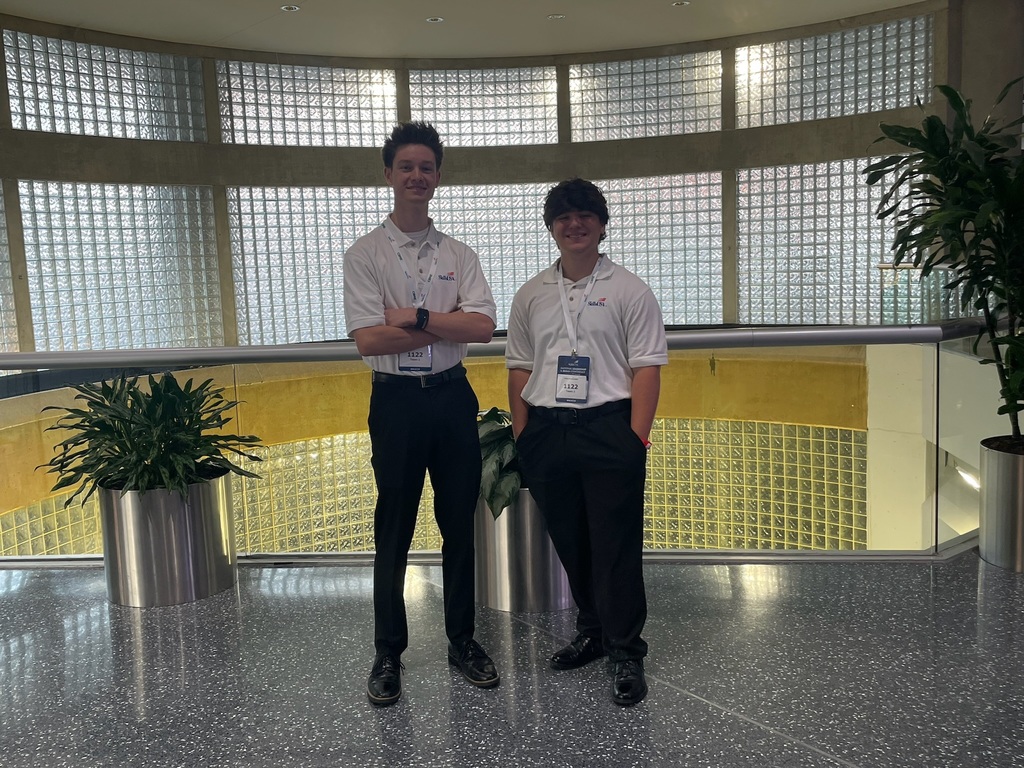 Two individuals wearing white shirts, black pants, and name tags stand indoors in front of a curved glass block wall. Large potted plants are positioned on either side of them, and the floor is made of a shiny, speckled material.