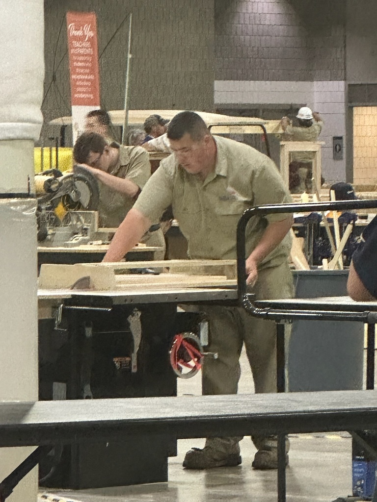 Several individuals in uniforms work in a workshop or industrial setting, engaged in woodworking activities using tools like saws and workbenches. A partially visible sign is in the background.