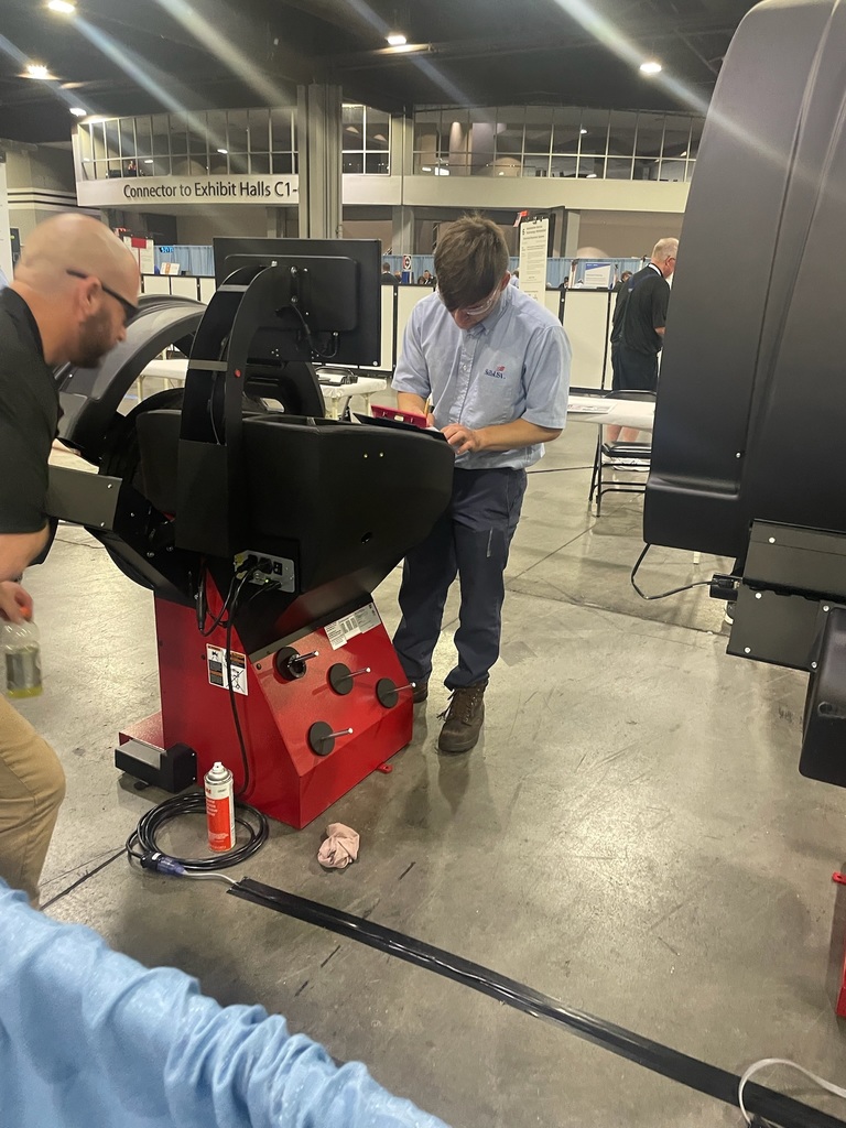 Two men work with a piece of industrial machinery in an exhibition hall. One man is bending over the machine, possibly making adjustments, while the other stands nearby writing or inspecting. The machine has a red base with knobs and buttons, and tools including a spray can and cloth are on the floor. Directional signs to exhibit halls are visible in the background.