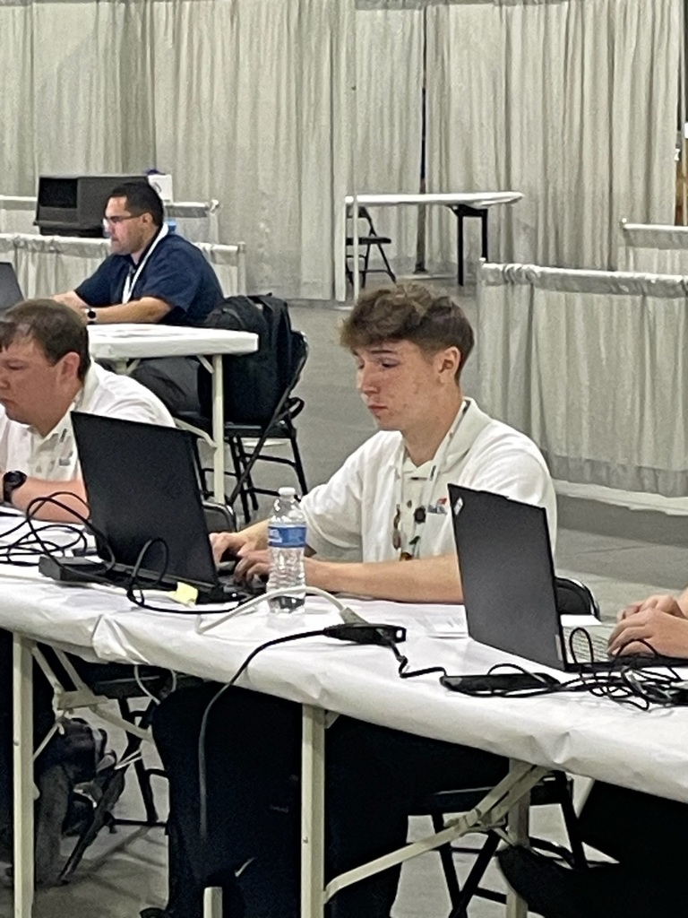 Several people are seated at tables with laptops in a conference or event setting. The background features partition curtains and additional tables.