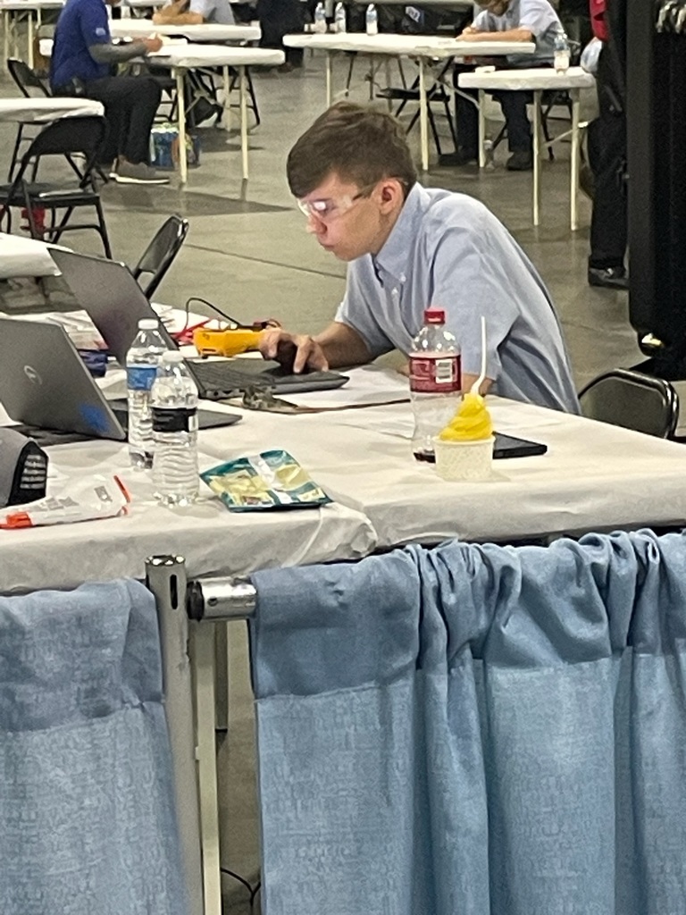 A person sits at a table with a laptop, surrounded by items including water bottles, a red drink bottle, a yellow dessert in a cup with a spoon, and papers or packets. Other individuals are visible in the background, also working at tables with laptops in an indoor event or workspace setting.