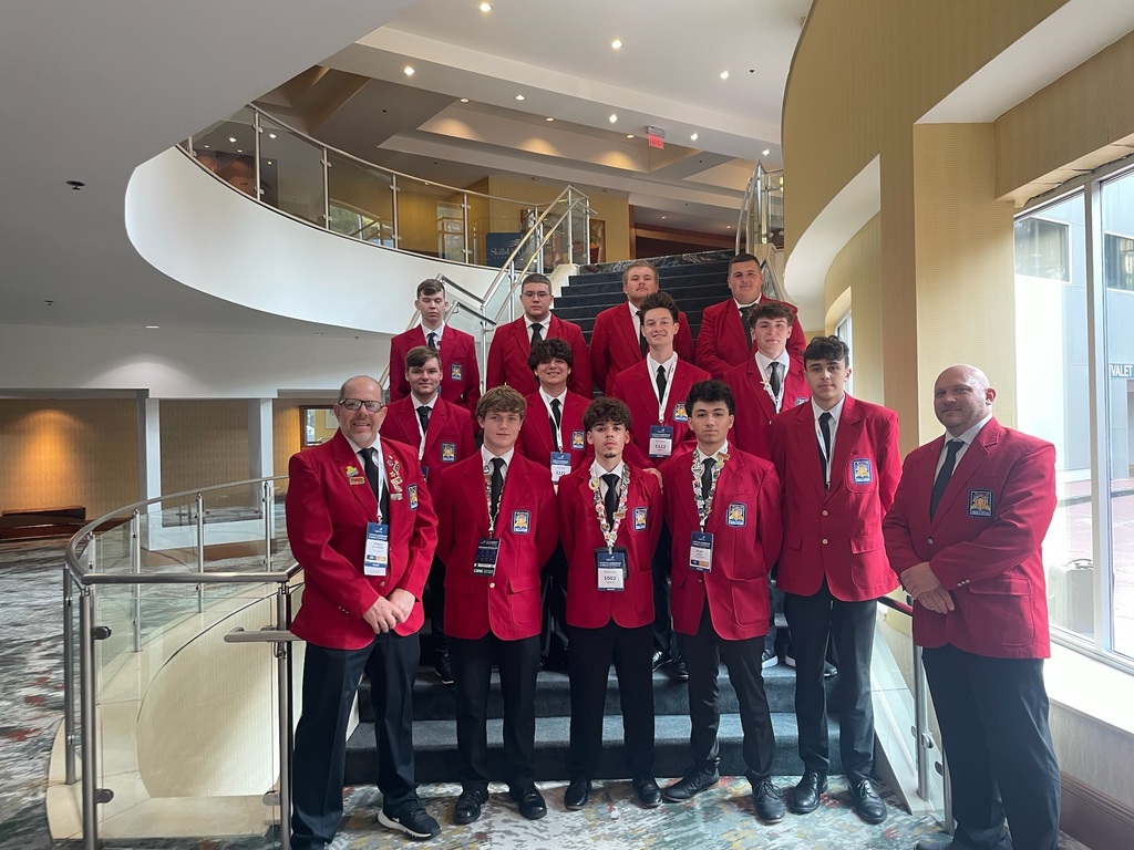 A group of individuals wearing red blazers with badges and name tags stand on a curved staircase inside a modern building. The staircase has glass railings, and the interior features contemporary design elements. The group appears to be part of an organization or attending an event.