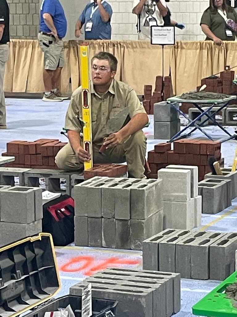 A person kneels on the ground holding a yellow level tool vertically, surrounded by construction materials such as cinder blocks and red bricks. Other individuals are in the background, observing or participating in what appears to be a masonry or construction demonstration.