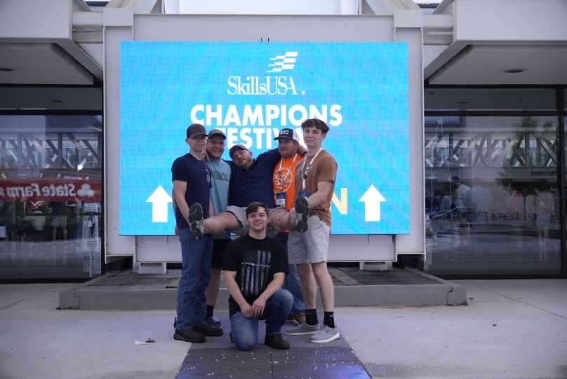 Five individuals pose in front of a large digital sign that reads 'SkillsUSA CHAMPIONS FESTIVAL' with two white arrows pointing upward. Four people are standing and holding a fifth person horizontally across them. Behind them are glass doors and windows reflecting the outdoor surroundings.