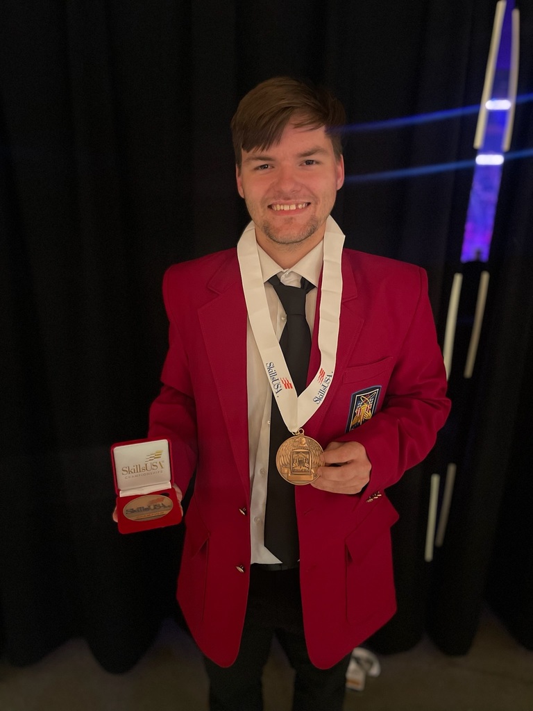 A person wearing a red blazer with a patch on the left side holds a medal in their right hand and an open box containing another medal in their left hand. They are also wearing a white 'SkillsUSA' lanyard around their neck with an additional medal attached. The background is dark with light streaks visible.