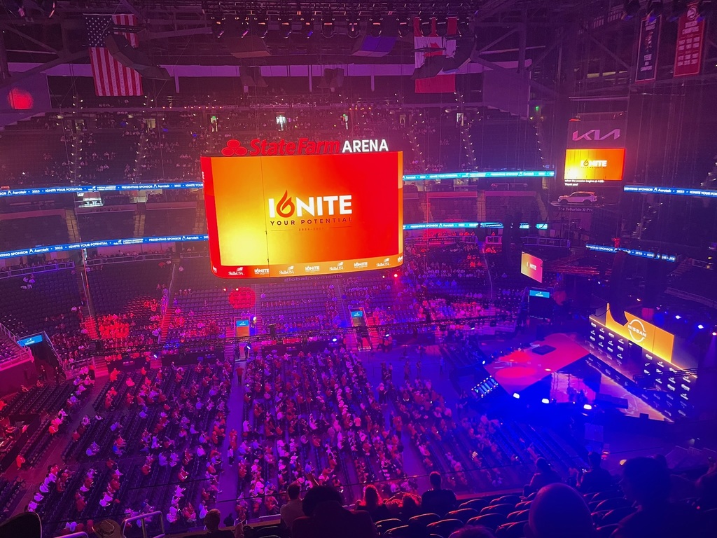 An indoor arena, identified as State Farm Arena, is filled with a large seated audience facing a central stage. A large screen displays the phrase 'IGNITE YOUR POTENTIAL' in bold letters against an orange and red background. Smaller screens around the arena show similar visuals. The lighting is primarily purple and blue, creating a vibrant atmosphere.