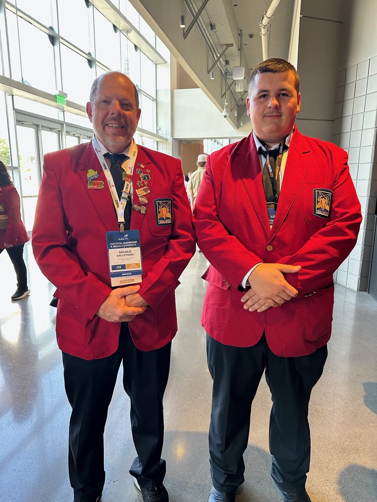 Two individuals stand side by side indoors, possibly in a convention center. Both are wearing red blazers with badges on the left chest. The person on the left has a name badge that reads 'SkillsUSA' and 'Advisor'.
