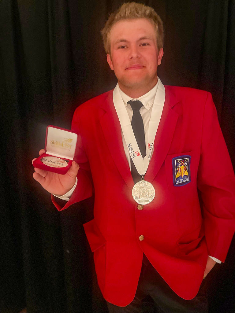 A person wearing a red blazer with a SkillsUSA patch, a white shirt, and a black tie holds an open box containing a medal or award in their left hand. A second medal hangs from a white 'SkillsUSA' lanyard around their neck. The background consists of dark, possibly black, curtains.