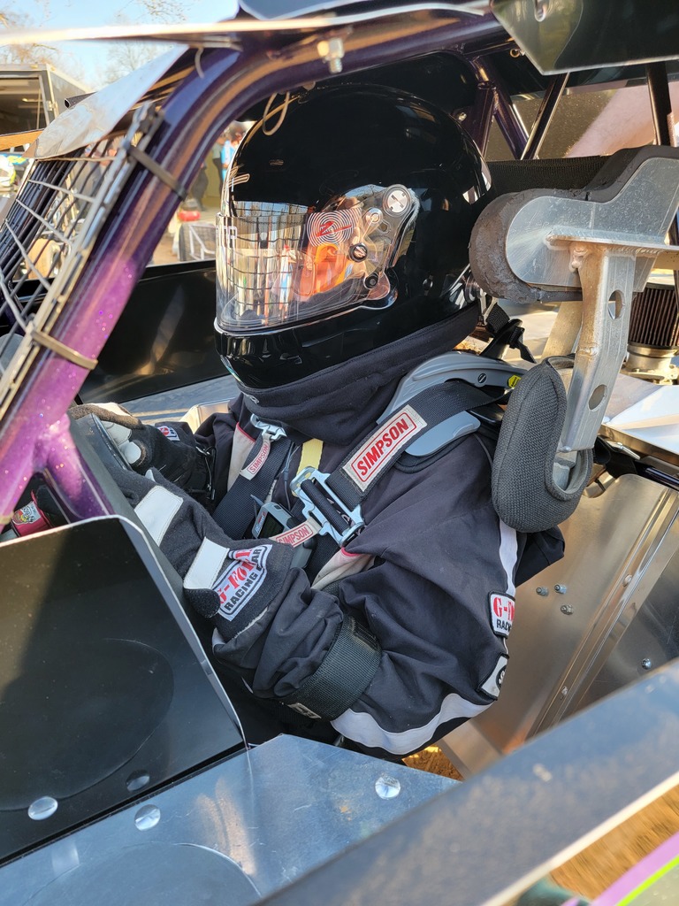 A race car driver is seated inside a race car, wearing a black helmet with a reflective visor and a black racing suit. The driver is secured with multiple safety harnesses labeled 'Simpson' and 'Impact Racing.' The interior of the car shows various metal components and parts of the roll cage. The image captures the driver from the side, focusing on the upper body and helmet.
