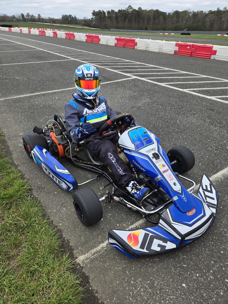 A person in a blue racing suit and helmet is seated in a blue go-kart on an asphalt track. The go-kart features various sponsor logos and the number 4 on the front. The track is marked with white lines and bordered by red-and-white barriers. Grass is visible on the left side of the image.