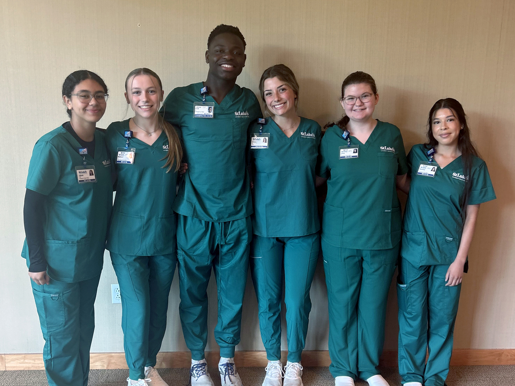A group of six individuals wearing green medical scrubs stands in a line against a beige wall. Each person has an identification badge clipped to their scrub top. The individuals have their faces blurred for privacy. They are standing close together with some having their arms around each other’s shoulders. The floor is carpeted, and there is a wooden baseboard along the bottom of the wall.