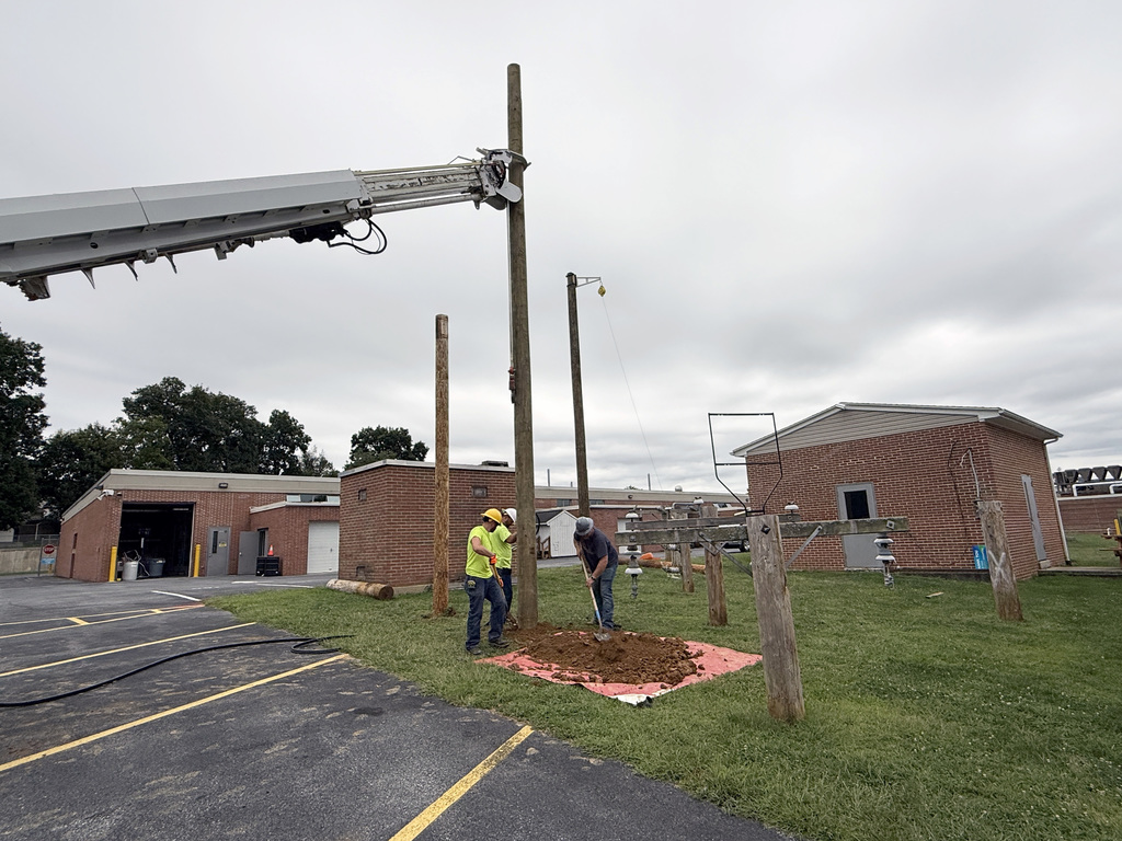 wo workers are installing a utility pole in a grassy area near a brick building. One worker wears a yellow safety vest and helmet, while the other wears casual work clothes. They stand on a red tarp surrounded by dirt at the base of the pole. A crane arm on the left assists with lifting and positioning. Several utility poles and related equipment are visible in the background under an overcast sky.