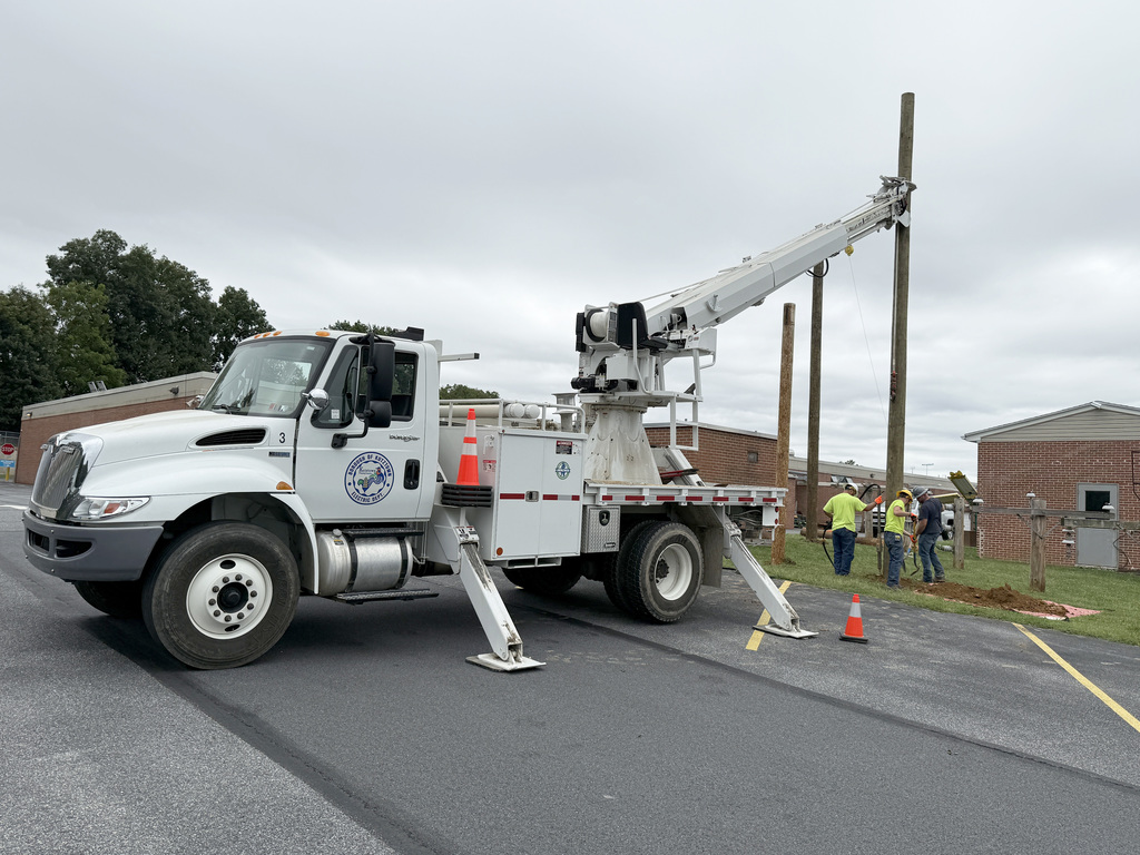 A white utility truck with a crane is parked on an asphalt surface. The crane is extended and being used to install or repair a wooden utility pole. Three workers in high-visibility clothing are standing near the base of the pole, which is situated on a grassy area next to a brick building. Orange traffic cones are placed around the work area for safety.
