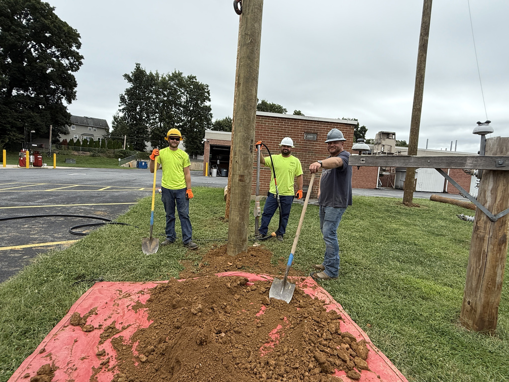 Three construction workers wearing helmets and high-visibility shirts are using shovels to move dirt onto a red tarp on a grassy area. They are working around a utility pole. In the background, a parking lot with yellow bollards and a brick building is visible.