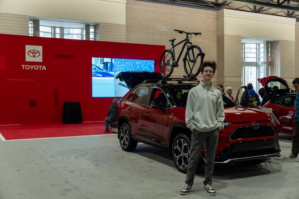 A red Toyota SUV is displayed at an indoor auto show. The vehicle has two bicycles mounted on the roof rack. A person in a white hoodie and gray pants stands in front of the car with hands in pockets. In the background, there is a red Toyota display booth with a large screen showing images and another person sitting on a chair.