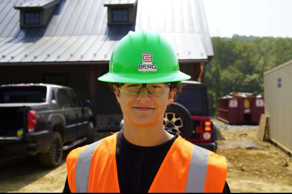 A person wearing a green hard hat labeled "BERG" and an orange safety vest stands at a construction site. The background features a black truck, a red Jeep, buildings, and trees. 