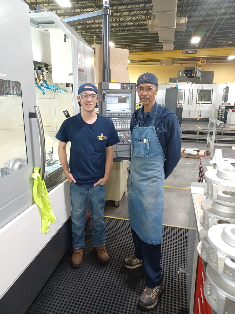 Two individuals stand in a workshop or factory setting. One wears a blue t-shirt, jeans, and a cap; the other wears a denim apron over dark clothing. They are positioned on a black rubber mat in front of industrial machinery, including CNC machines and various tools and components.