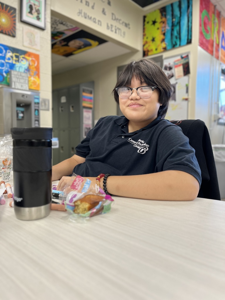 A person sits at a table in a classroom or office setting, wearing a dark-colored polo shirt with "The Center School" and the number "13" on it. Their face is blurred for privacy. On the table are a black travel mug, snacks, and other small items. The background features colorful posters, decorations, lockers, and classroom materials.