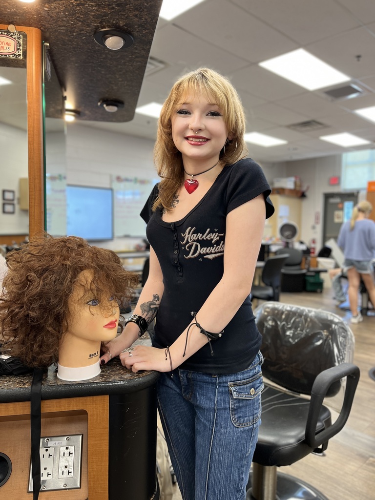 A person wearing a black "Harley-Davidson" t-shirt and jeans stands next to a mannequin head with curly hair on a counter in a classroom or training room, possibly for cosmetology. Chairs, desks, and other people are visible in the background.