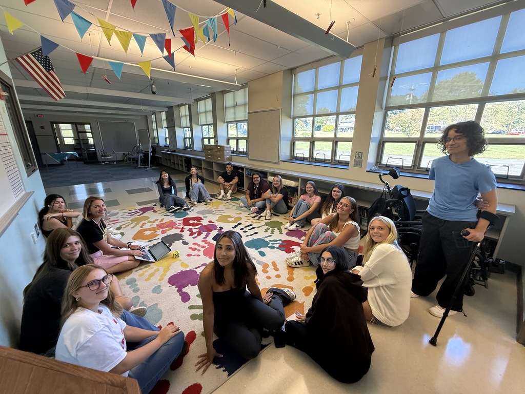 A group of people sit in a circle on a colorful rug inside a classroom. The room features large windows, an American flag, and colorful triangular banners hanging from the ceiling.