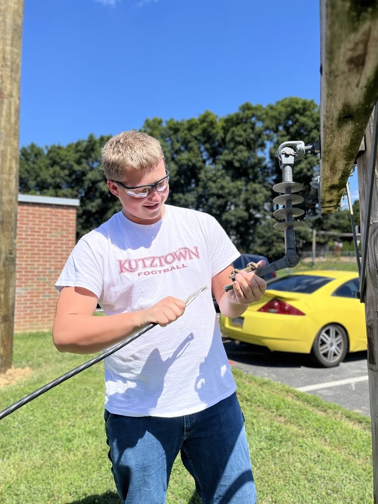 A person wearing a white "Kutztown Football" t-shirt and blue jeans is working with electrical equipment outdoors. They are holding a tool and appear to be adjusting or installing a component on an electrical pole. In the background, a yellow car is parked on the grass near trees and a brick building.