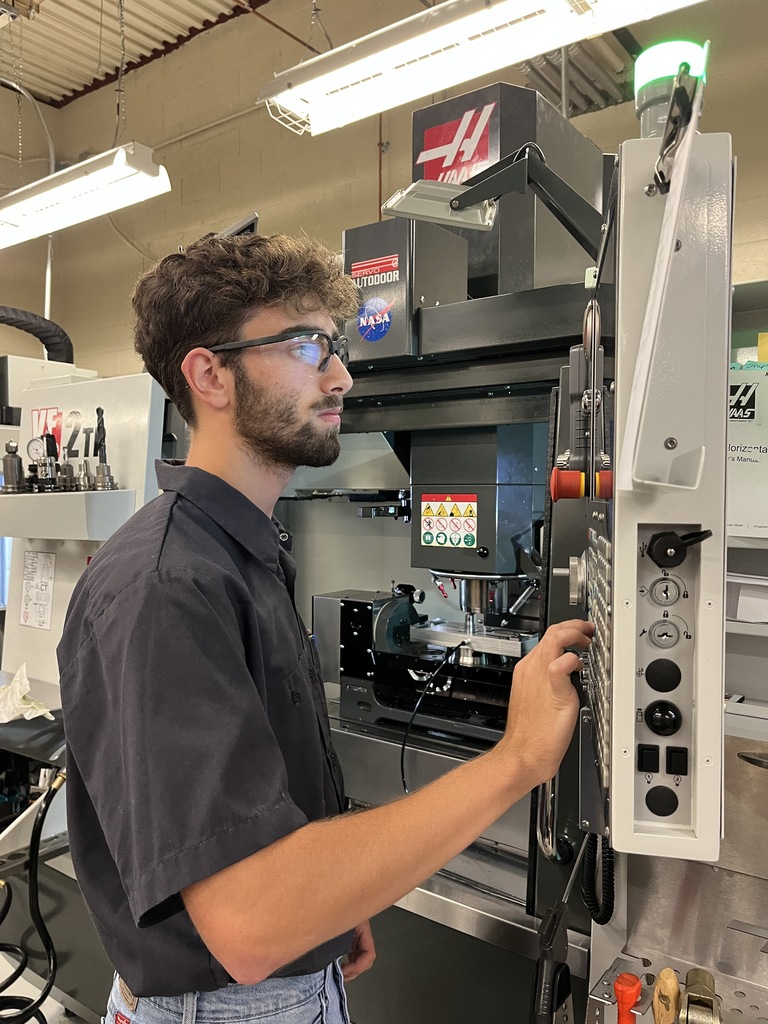 A person with a blurred face is operating a Haas CNC machine in an industrial setting. The person is wearing a dark shirt and jeans, and is focused on the control panel of the machine. The background shows various parts of the CNC machine and fluorescent lights hanging from the ceiling.