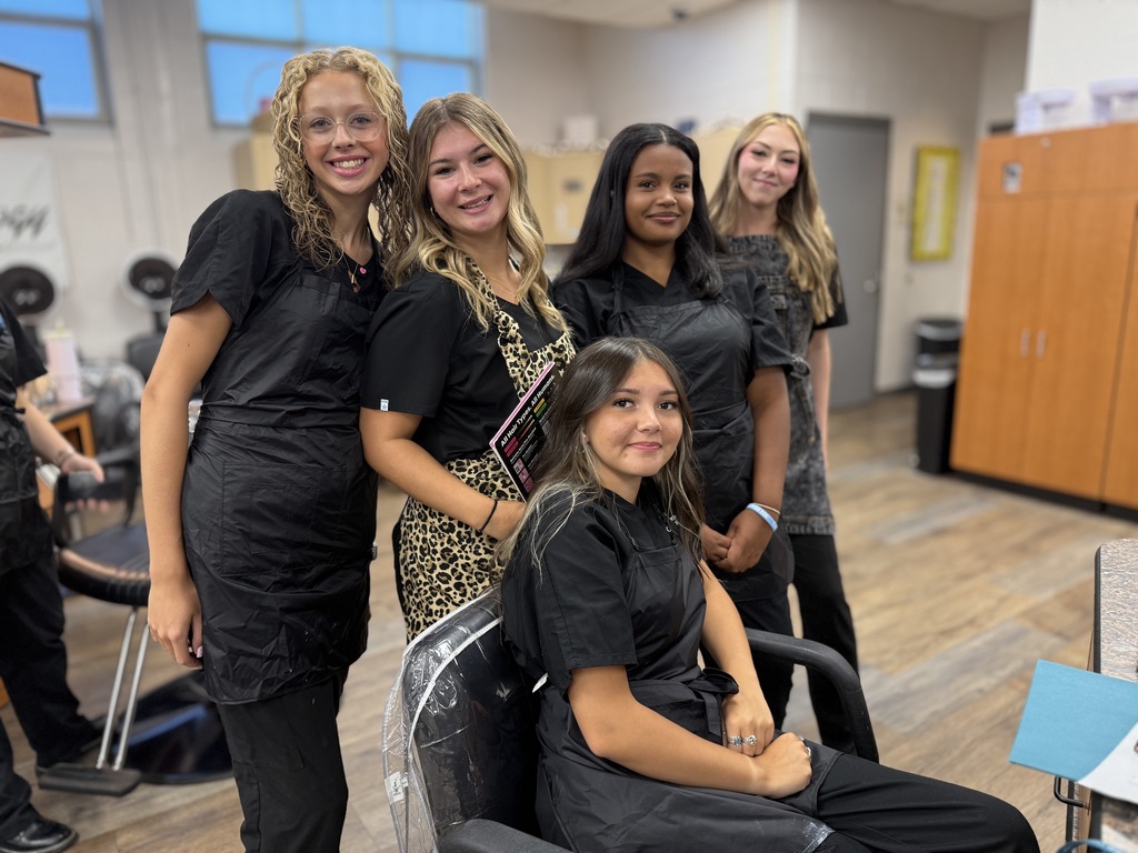 Five individuals are in a salon or cosmetology classroom. Four are standing, and one is seated in a salon chair. All are wearing black outfits, with some in aprons. The background includes salon equipment such as chairs and cabinets.