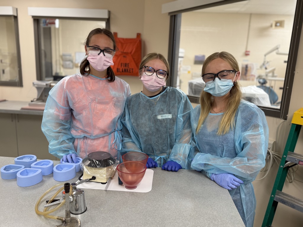 hree individuals wearing protective gowns, gloves, and face masks stand in a laboratory setting behind a counter. The counter holds various laboratory equipment, including blue molds, a vacuum pump with tubing, and two bowls.