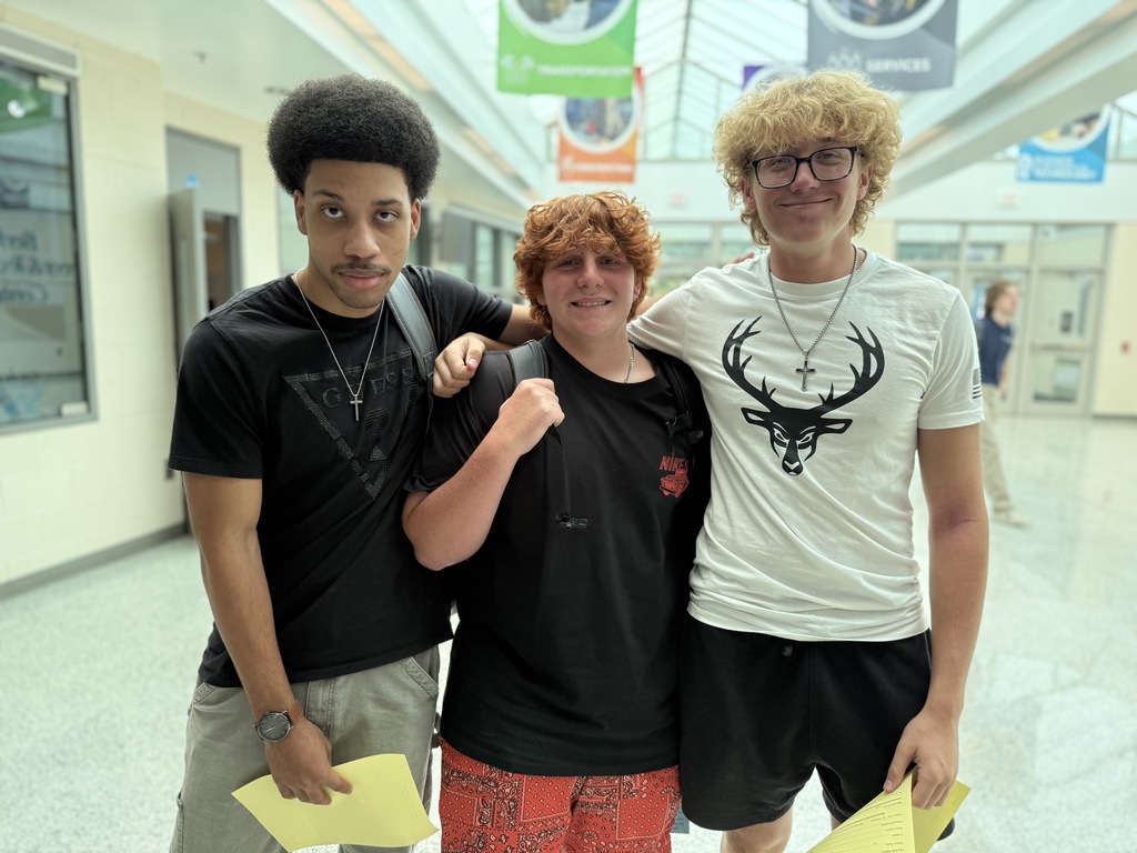 Three individuals stand close together in a hallway, each holding a yellow piece of paper. Their faces are blurred for privacy. The person on the left wears a black shirt and gray shorts, the middle person wears a black shirt and red patterned shorts, and the person on the right wears a white shirt with a deer head design and black shorts. The hallway features banners hanging from the ceiling and doors along both sides.