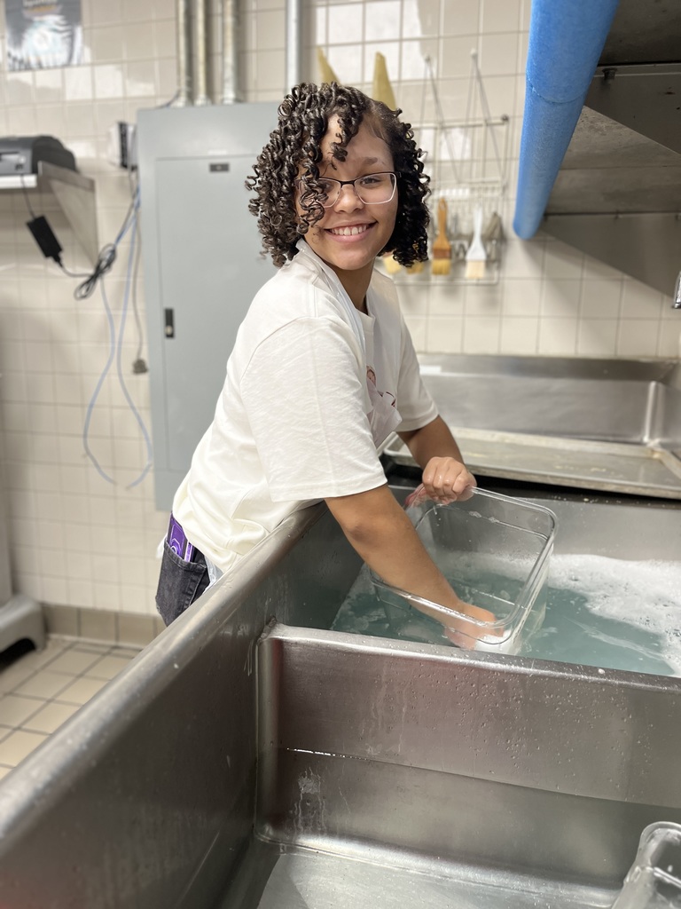 A person is washing a rectangular glass container in a large stainless steel sink filled with soapy water. The person is wearing a white t-shirt. The background features tiled walls, an electrical panel, and various kitchen utensils hanging on the wall.