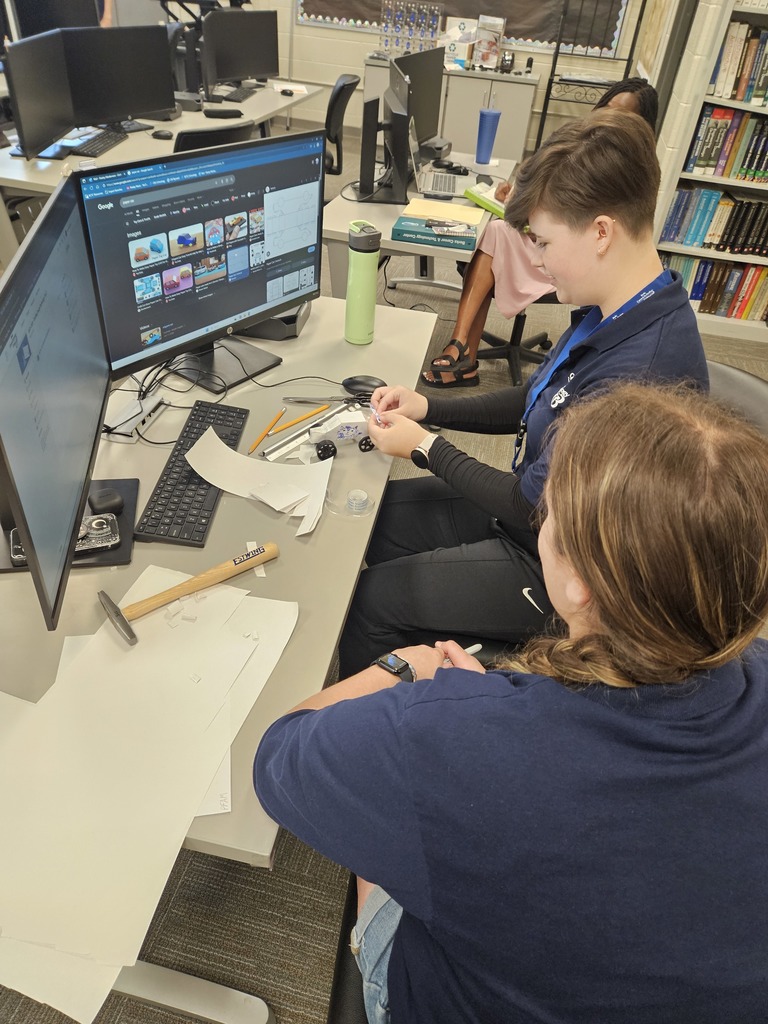 Two individuals sit at a desk in a classroom or lab setting, collaborating on a project. The desk contains papers, a hammer, pens, and a computer monitor displaying a platform with multiple video or image thumbnails. One person holds an object while the other observes, indicating active engagement in a hands-on learning or problem-solving activity.