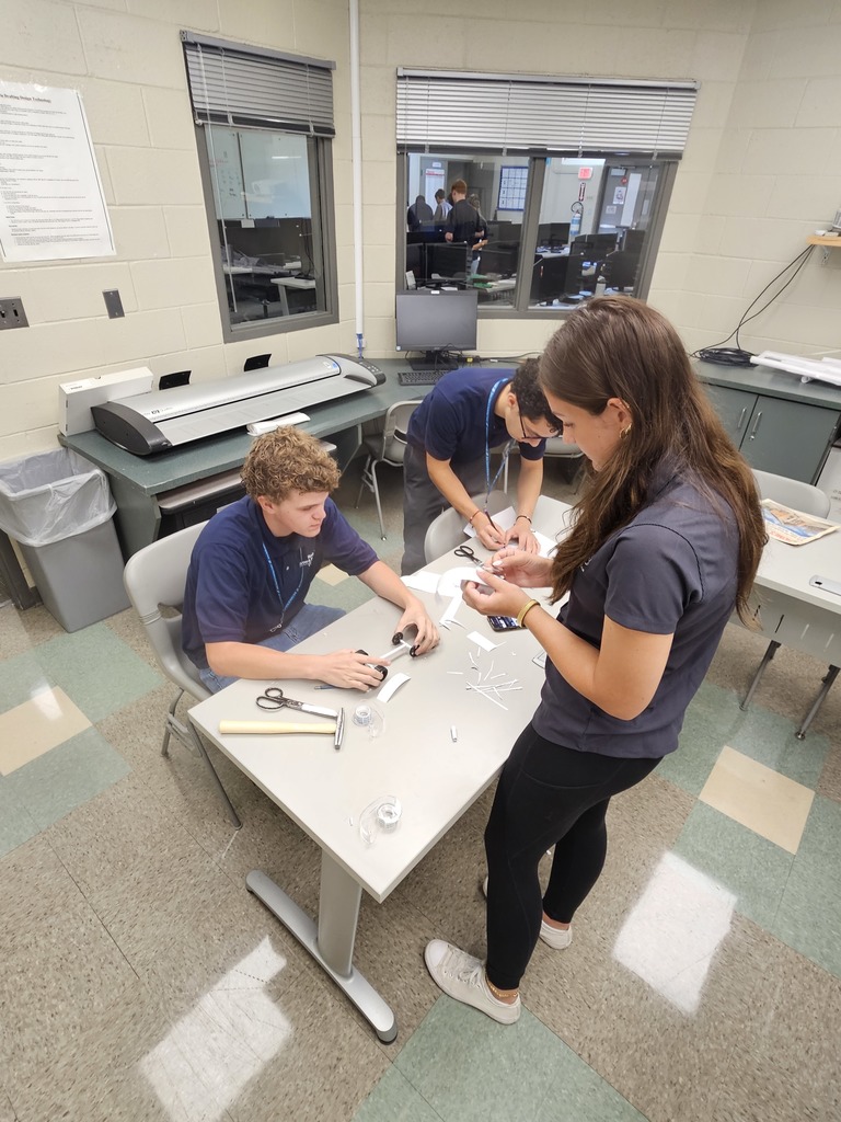 Three individuals collaborate at a table in a classroom or lab setting, working with small white sticks, a ruler, and other tools. One person stands while two are seated, focused on the task. The background includes office equipment like printers and computers, and windows reveal another room with additional people engaged in work, emphasizing a hands-on, team-oriented environment.
