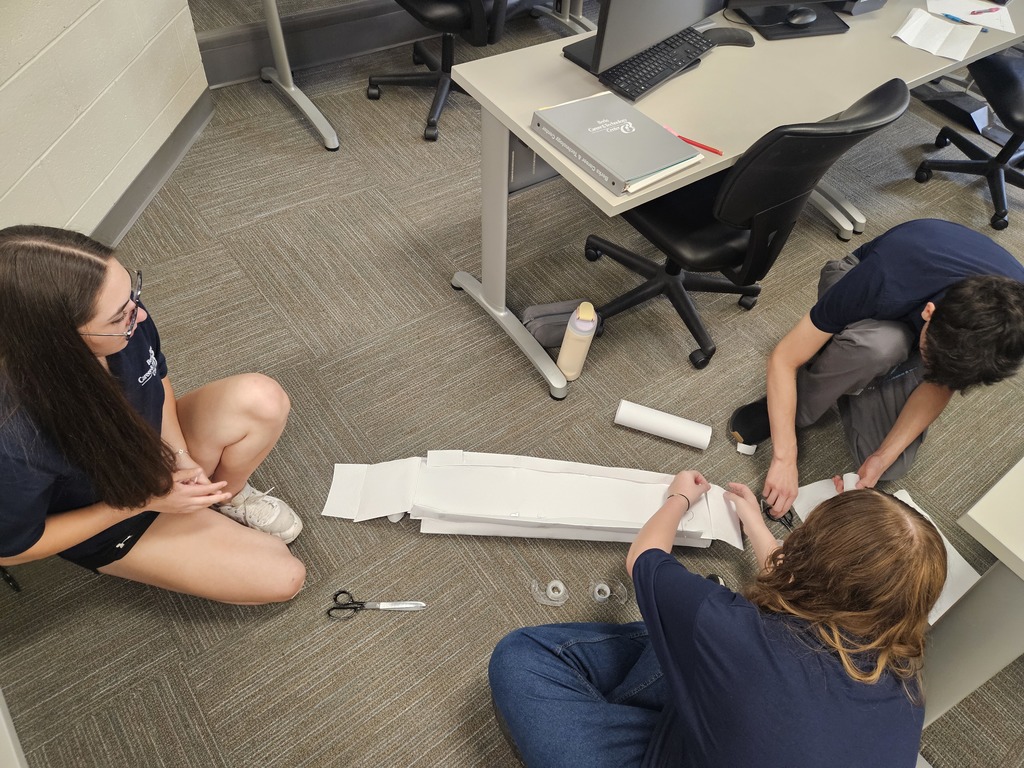 Three individuals sit on the floor in a collaborative workspace, surrounded by desks and chairs. They are engaged in a hands-on project using white paper or cardboard, scissors, and tape. A nearby desk holds a laptop, notebook, and water bottle, indicating an active and creative work session in a classroom or office setting.