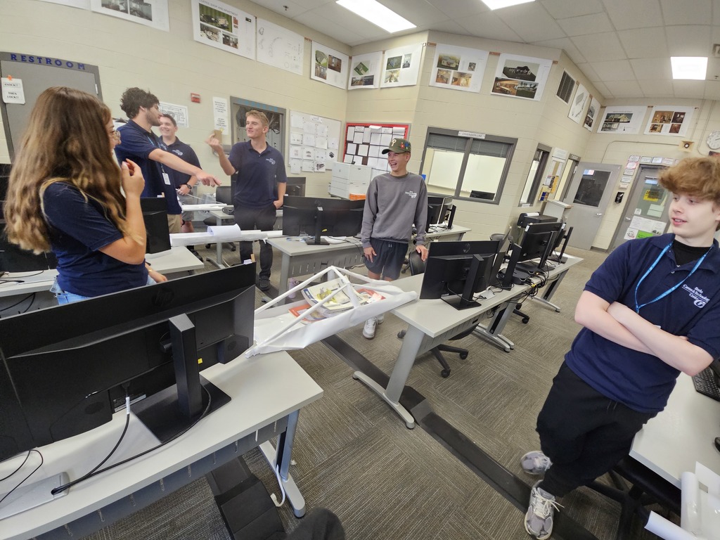 Several individuals stand and interact around computer desks in a classroom equipped with desktop computers. A white model structure, possibly related to engineering or architecture, is placed on one of the desks. The walls feature posters and images, suggesting an educational setting focused on collaborative learning and project-based activities.