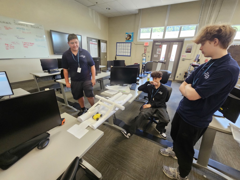 Three individuals in matching dark blue shirts work together in a classroom or lab setting. They are focused on a small structure made of white pipes and a yellow toy car, possibly part of a hands-on experiment or project. The room features computer workstations, monitors, and a whiteboard with partially visible writing, suggesting an educational or training environment.