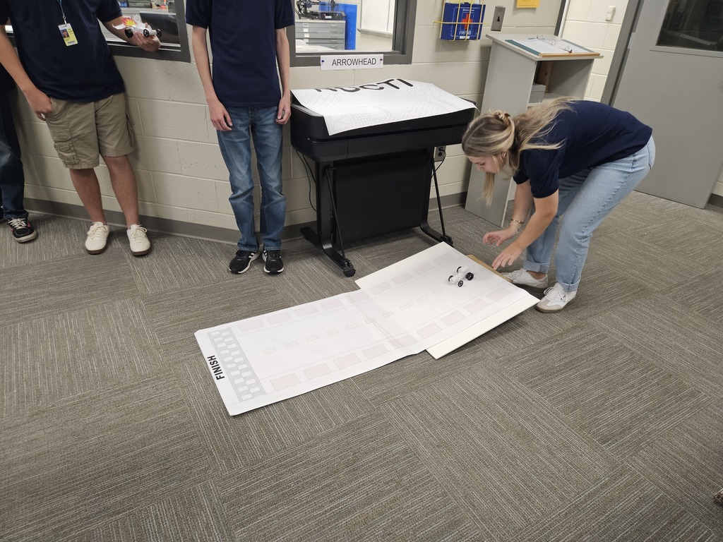 In a room with grey carpet and light-colored walls, one individual kneels on the floor adjusting two small wheeled robots on a large sheet of paper marked with a track and the word "FINISH." Three others stand nearby observing. A large printer labeled "ARKANSAS EC" is visible in the background, suggesting a robotics-focused educational or project-based activity.
