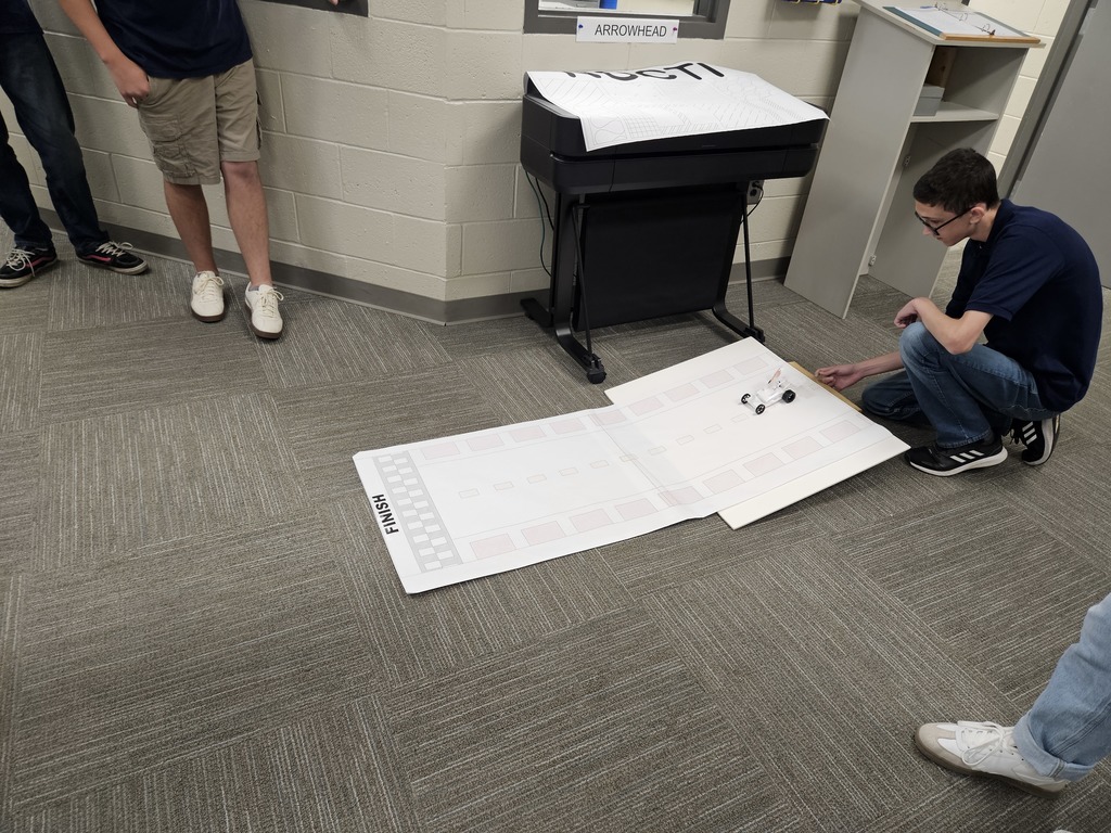 n a room with gray carpet and light-colored walls, one individual kneels beside a large sheet of paper marked with a track and the word "FINISH." Small objects resembling miniature vehicles or robots are placed on the track. Other individuals stand nearby, observing the setup. The environment suggests a robotics or engineering activity in an educational or project-based setting.