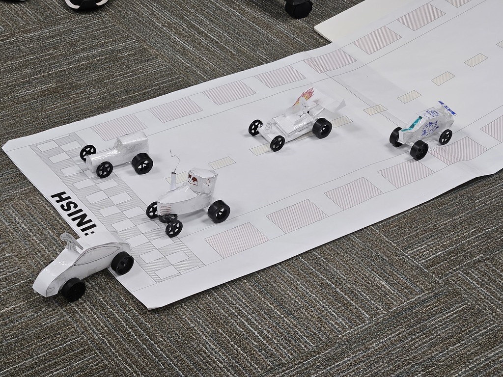 A small-scale race track is set up on a carpeted floor, featuring five miniature cars made from lightweight materials. The track includes marked lanes and the word "HSINI" printed at one end, suggesting a school project or competition focused on engineering, design, or creativity.