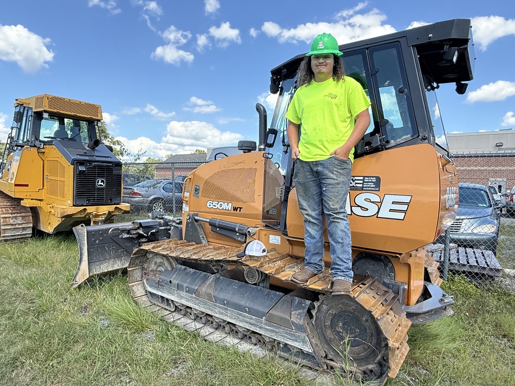 A person wearing a green hard hat and a bright yellow-green safety shirt stands on the track of an orange CASE 650M WT bulldozer. The person has one hand in their pocket and is smiling. In the background, there is another yellow bulldozer, several parked cars, and a building with a brick facade. The sky is blue with scattered clouds.